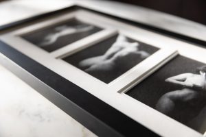 A close-up of a framed prints for boudoir photos display, featuring three black-and-white boudoir images arranged in a triptych-style frame. The prints are matted with a white border, enclosed within a sleek black frame that contrasts with the soft, intimate imagery inside. The photographs depict the female form in an artistic, blurred style, emphasizing curves and movement through delicate lighting and shadow play. The frame rests on a marble surface, with natural light casting subtle reflections on the glass. The composition highlights the elegance of fine art boudoir photography, capturing the sensuality and sophistication of the printed images.