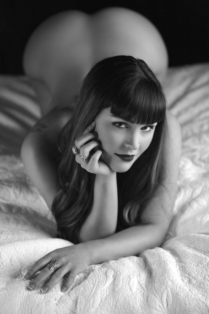 A black-and-white boudoir portrait of a woman lying on a bed, gazing directly into the camera with a confident, playful expression. Her dark eyes are framed by bold eyeliner and soft waves of hair, and her slightly parted lips hint at a knowing smile. One hand rests gently on her cheek, adorned with ornate rings that add elegance to the composition. The textured quilt beneath her contrasts with the smoothness of her skin, while the blurred background emphasizes graceful lines and feminine form. Soft lighting adds intimacy and depth, showcasing the artistry and empowerment captured in professional boudoir photography.