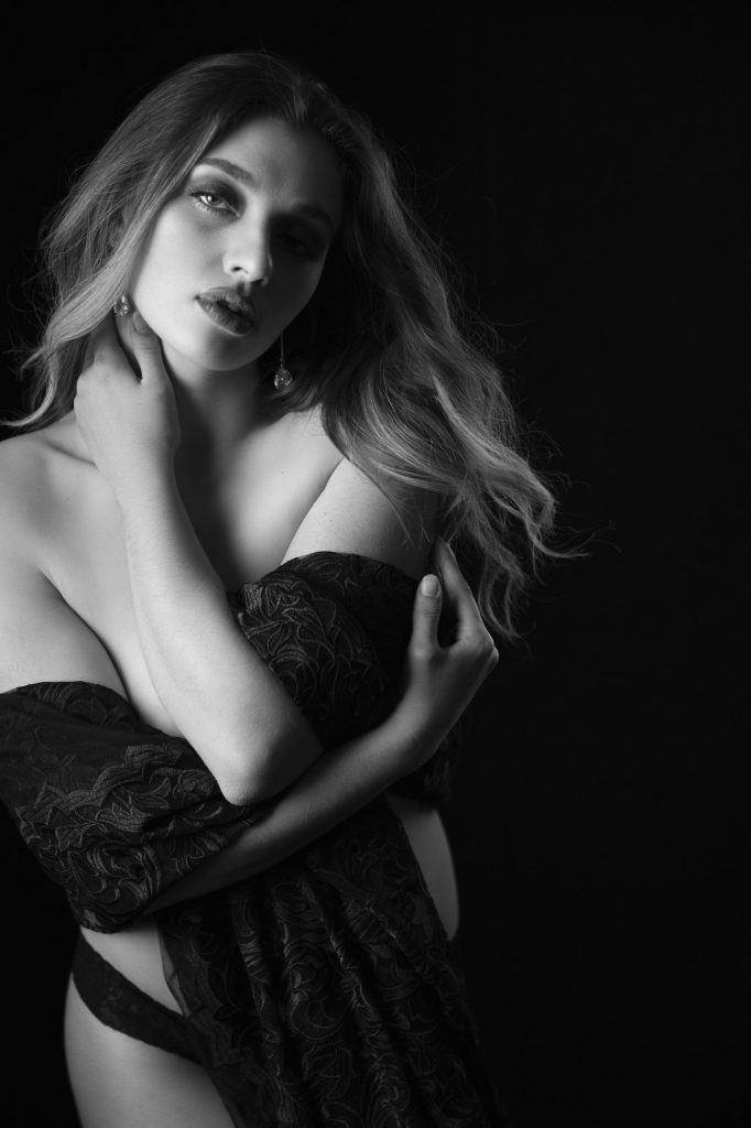 A black-and-white, magazine-style boudoir photography portrait featuring a woman draped in patterned black lace. She gazes intensely into the camera, her parted lips and dramatic makeup adding to the editorial mood. Her wavy hair flows over her shoulders, while soft shadows sculpt her cheekbones, collarbone, and lips. The pose, with one hand on her neck and the other across her torso, evokes both vulnerability and strength. Shot against a minimalist dark background, the image blends fashion, intimacy, and fine art in a timeless composition.