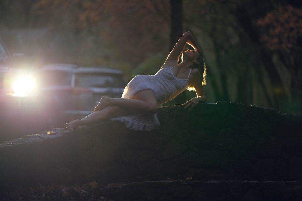 A moody outdoor boudoir portrait captures a woman reclining on a rustic stone wall, backlit dramatically. She wears a delicate white lace slip that contrasts softly with the rugged stone beneath her. Her relaxed and expressive pose includes one arm draped over her head and the other supporting her weight as she tilts her face toward the fading light. The golden glow of a car’s headlights illuminates the misty evening air, creating a cinematic atmosphere. Silhouettes of trees and parked vehicles fade into soft darkness in the background, adding depth and mystery. Wisps of hair catch the light, framing her face with an ethereal glow. The contrast of shadow and illumination enhances the artistry and mood of this outdoor boudoir session.