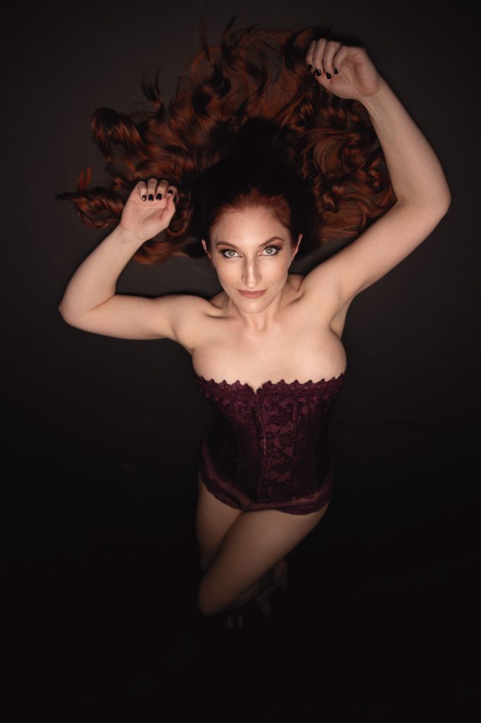 A stunning woman with fiery red hair lies against a dark background, her cascading curls fanning around her. She wears a deep burgundy corset with delicate lace details that accentuate her hourglass figure. Her piercing green eyes lock onto the camera, exuding confidence. Dramatic overhead lighting casts soft shadows, emphasizing her toned arms and décolletage. Her hands rest elegantly near her head, with black-painted nails adding bold sophistication. This artistic image reflects the work of a Santa Rosa boudoir photographer blending classic elegance with modern style.