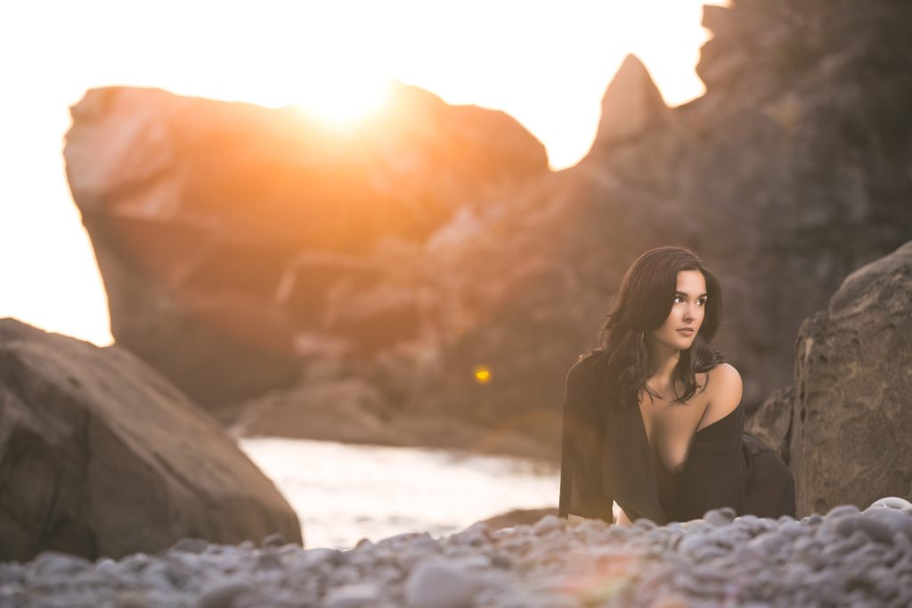 A woman with dark, wavy hair kneels on a rocky shoreline at golden hour, wearing a loosely draped black robe that reveals a graceful silhouette. She gazes thoughtfully into the distance as warm sunset light illuminates her skin and highlights the textures of the coastal cliffs and shimmering water. The soft glow and natural surroundings create a dramatic, artistic atmosphere, blending feminine strength with serene beauty in a refined beach portrait.