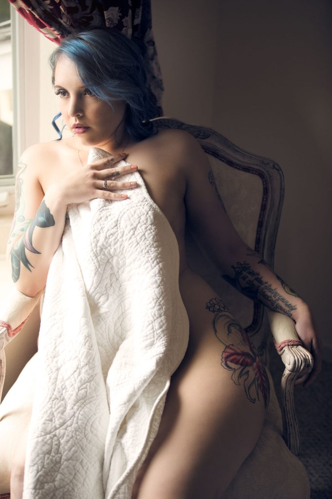 A tasteful boudoir portrait shows a woman draped in a delicate white quilt, seated in an ornate vintage chair. She gazes thoughtfully toward the window, with soft natural light highlighting her fair skin and intricate tattoos. Her blue hair cascades in soft waves, contrasting beautifully with the warm, muted tones of the room. The quilt subtly conceals her form while offering glimpses of her curves. Her hand rests gently on her chest, adorned with rings, adding a personal touch. The dreamy lighting and classic furnishings create an atmosphere of elegance, confidence, and intimacy.