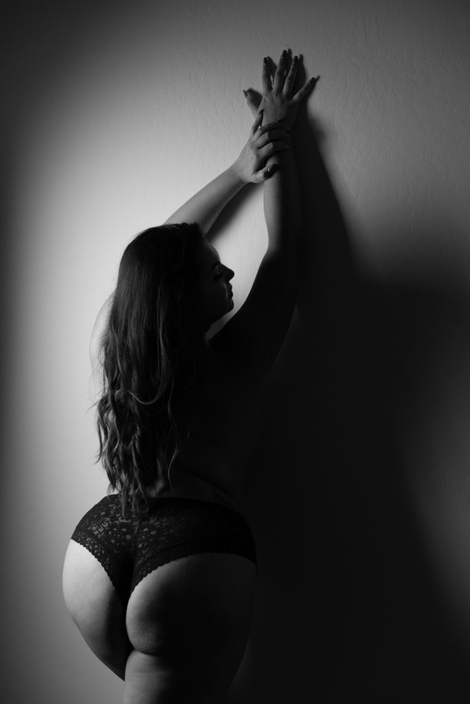 A black and white boudoir portrait features a plus-size woman gracefully posed against a textured wall. Her arms are elegantly raised above her head, fingers gently touching as she gazes thoughtfully to the side. Dramatic lighting creates deep shadows that highlight the curves of her body and the soft waves of her long hair cascading down her back. She wears black lace lingerie that contrasts with the illuminated wall. The interplay of light and shadow emphasizes her confidence and natural beauty, reflecting empowerment and strength in this artistic boudoir image.