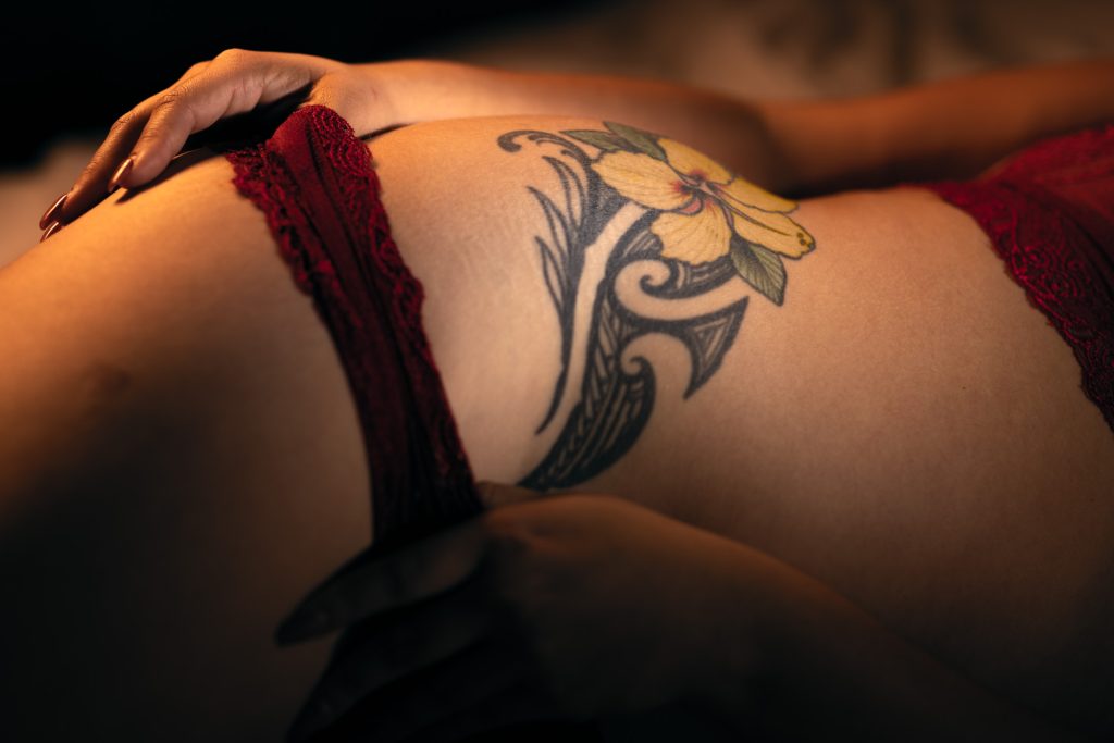 A close-up boudoir photograph focuses on the curve of a woman’s hip adorned with a detailed tattoo of a yellow hibiscus flower intertwined with bold black tribal patterns. Deep red lace lingerie contrasts against her smooth skin, with her fingertips gently resting on the fabric. Warm, moody lighting casts soft shadows, highlighting the intricate tattoo details and the texture of the lace. The composition emphasizes the artistry of the body and ink, capturing refined detail in boudoir photography.