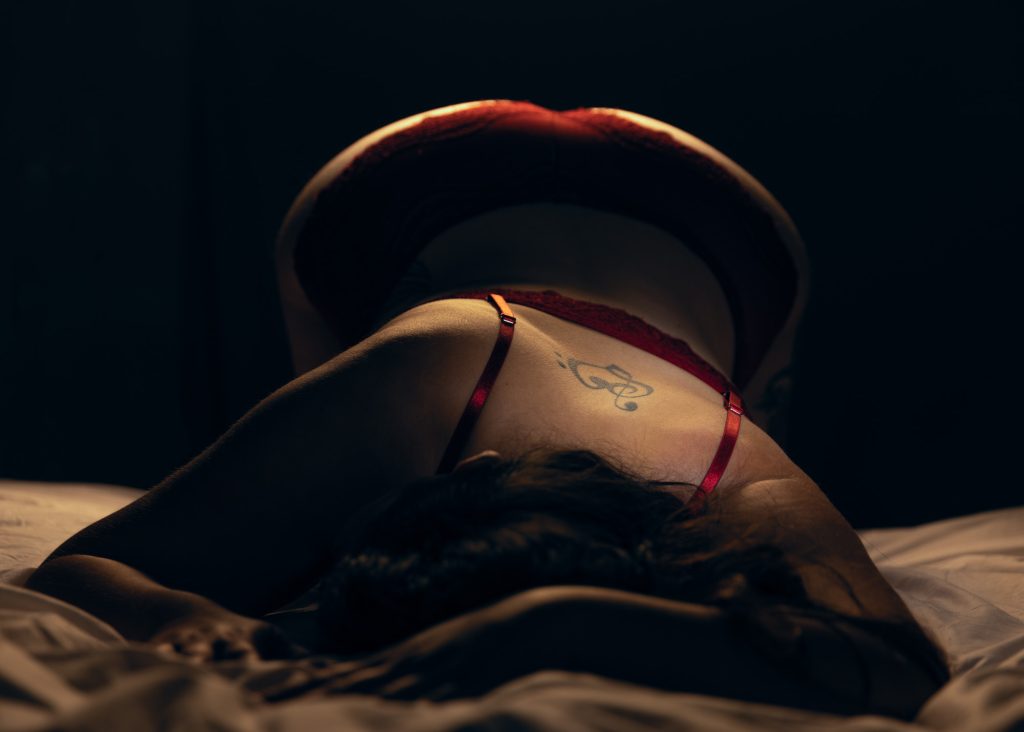 A dramatic boudoir portrait captures a woman posed on a bed, arching her back while wearing deep red lace lingerie. Soft lighting highlights the curves of her body, casting shadows that enhance the texture of the lace and the contours of her form. A delicate treble clef tattoo is visible on her upper back, adding a personal artistic detail. The red bra straps contrast with her smooth skin, and her long dark hair cascades over the sheets. The moody lighting and intimate pose create an evocative atmosphere, emphasizing the elegance of lace and form in boudoir photography.