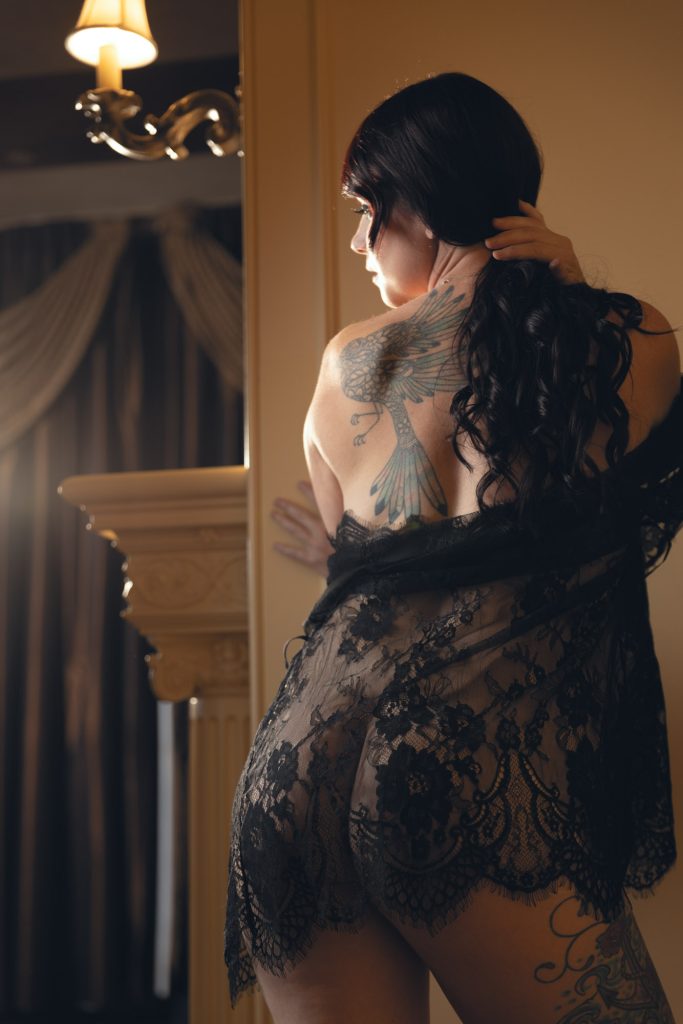 A luxurious high-end boudoir photography session in Sonoma County features a woman with long, dark curls cascading over her shoulder. She stands with her back to the camera, showcasing an intricate bird tattoo spanning her shoulder blade. A sheer black lace robe drapes over her curves, partially revealing her tattooed thigh. The warm glow of a vintage wall sconce casts soft light on her skin, while elegant drapery and a gilded column in the background enhance the opulent atmosphere. Her hand gently gathers her hair, adding a personal touch to the composition. The refined setting and expert lighting embody the elegance of high-end boudoir photography in Sonoma County.