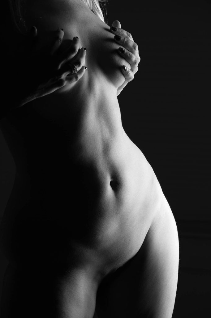 A black-and-white high-end boudoir photograph features a woman's torso in dramatic lighting. Her hands gently cradle her body, adorned with dark nail polish and elegant rings, adding a touch of sophistication. The interplay of light and shadow accentuates the graceful curves of her figure, highlighting the smooth contours of her waist and the delicate dip of her navel. The background fades into deep black, enhancing the contrast and creating a timeless, artistic composition. The image embodies luxury, elegance, and refined artistry in boudoir photography.
