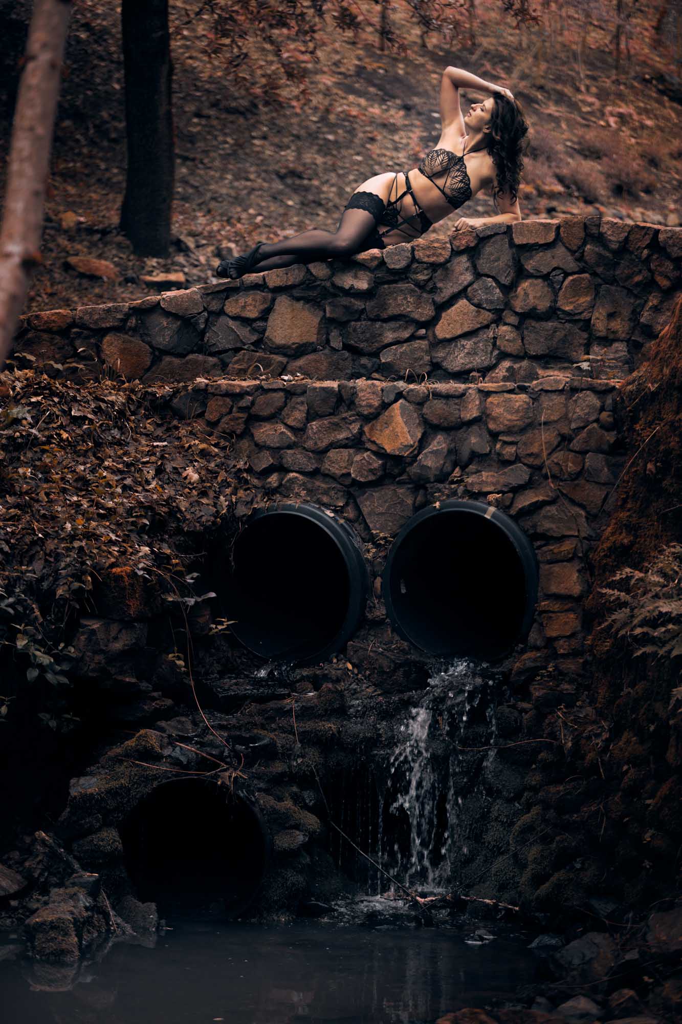 A woman with dark wavy hair poses on a stone wall for some tasteful outdoor boudoir photos in Santa Rosa. She wears a black lace lingerie set with stockings and high heels while one hand rests gently in her hair and her head tilts back. Soft natural light illuminates the wooded background with fallen leaves and water flowing from pipes below the wall.