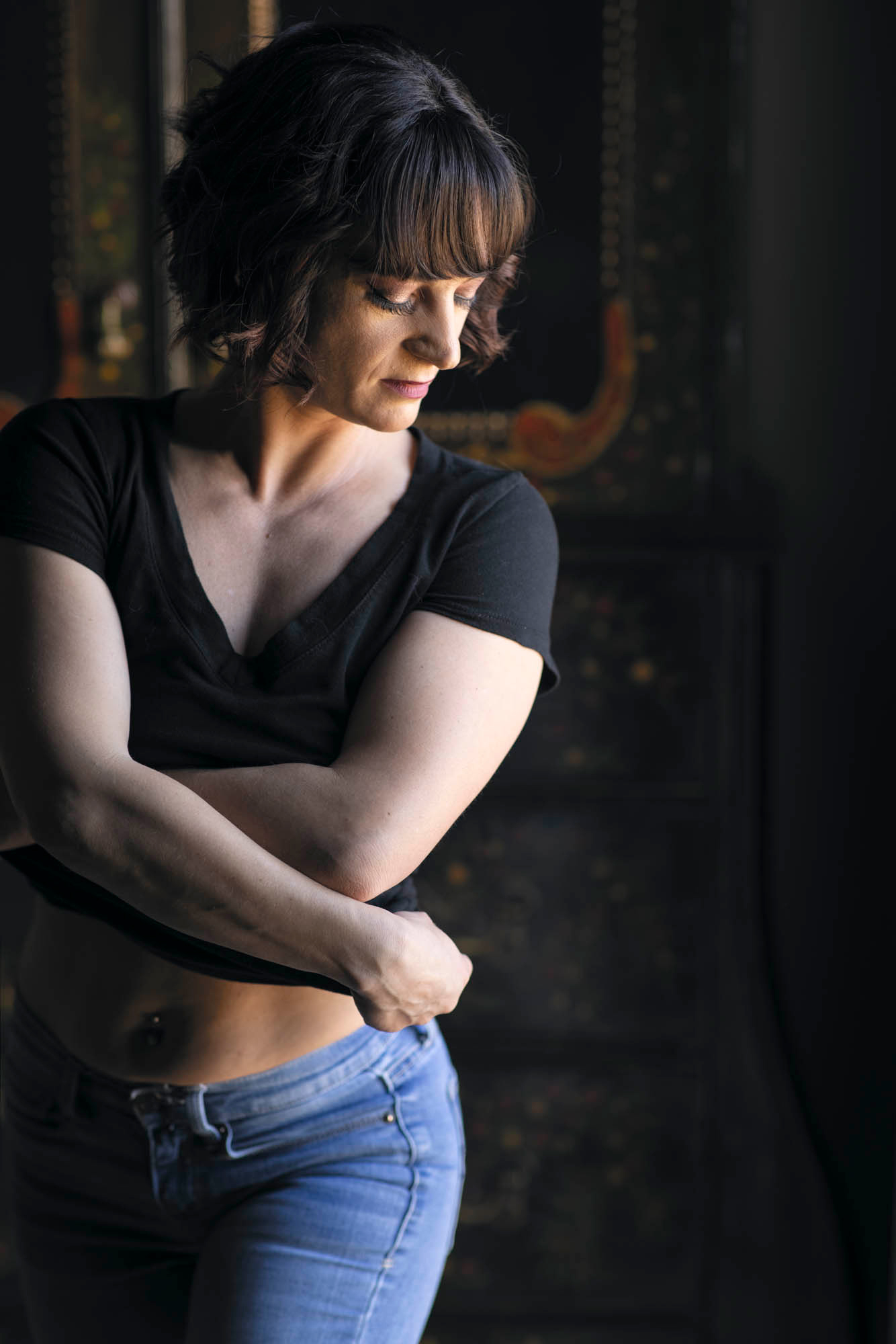 an with short dark hair and bangs crosses her arms over her chest in dramatic side lighting as she gazes downward during her empowering portraits Sonoma County session. She wears a black V-neck crop top and blue jeans against a dark ornate wooden background.