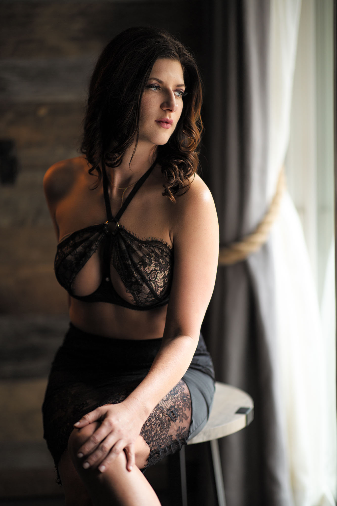 A woman with dark wavy hair poses for her lingerie photo shoot for every day women. In Santa Rosa photo shoot she sits on a wooden stool wearing a black lace halter top and matching skirt with her hands resting on her knee as she gazes to the side. Soft natural light from a window with curtains illuminates her against a rustic wooden wall.