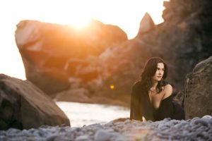 A woman with long dark wavy hair poses for her outdoor boudoir photography session. Dramatic rock formations rise behind her bathed in warm golden sunset light. The woman kneels on the Sonoma County pebble beach near the water wearing a black off shoulder garment and gazes to the side.