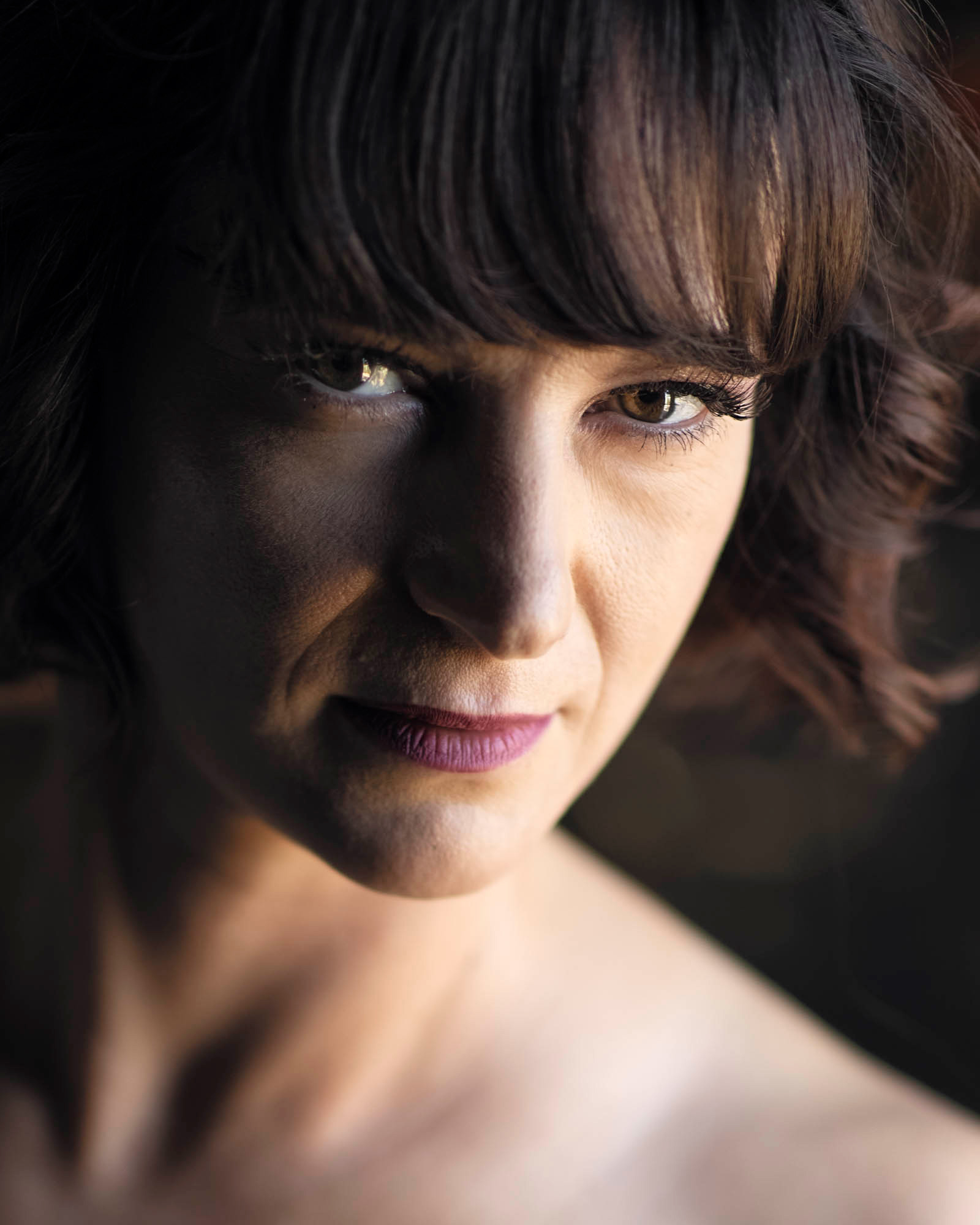 A woman with dark wavy hair and straight bangs poses for her sexy and confident portraits session. Dramatic side lighting illuminates her brown eyes and warm skin tones as she looks directly at the camera with a subtle smile. Her bare shoulders appear in the tight close-up composition while soft shadows add depth across her face.