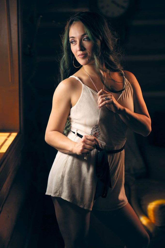 A woman with long, wavy dark hair accented with green highlights stands in a warmly lit, intimate setting. She wears a soft, off-white romper with thin straps, cinched at the waist with a dark sash. Her expression is gentle and confident as she gazes to the side, illuminated by the golden glow of light streaming through a nearby window. Large hoop earrings frame her face, and she delicately holds a long, intricate pendant necklace between her fingers, showcasing a subtle ring on her hand. The dimly lit background features wooden walls and a vintage-style clock, enhancing the cozy, rustic atmosphere. The soft shadows and warm tones add depth and emotion, perfectly embodying the essence of intimate portrait photography Sebastopol.