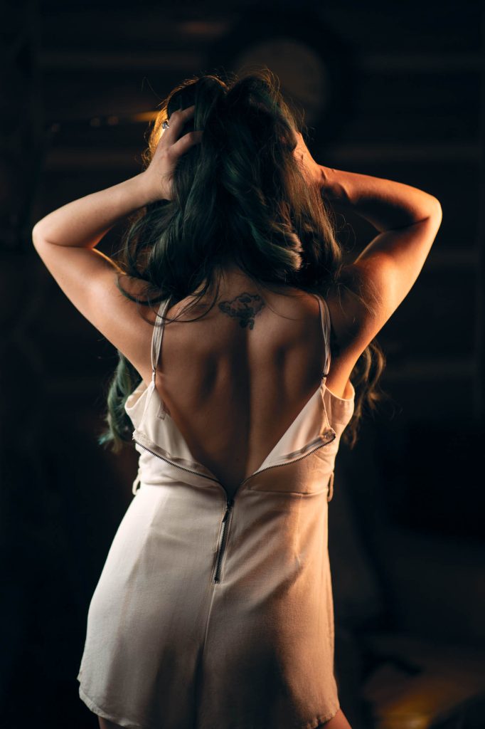 A woman stands with her back to the camera, her hands gently running through her long, wavy dark hair, which catches hints of green in the warm, moody lighting. She wears a loose, off-white romper with delicate straps slipping down her shoulders, exposing the curve of her back and a small, intricate tattoo just below her neckline. The partially unzipped back of the romper adds an effortless, undone elegance to the image. The dimly lit background, with rustic wooden walls and a vintage clock, enhances the dreamy, intimate feel of this boho style boudoir portrait, blending natural beauty with a carefree, sensual essence.