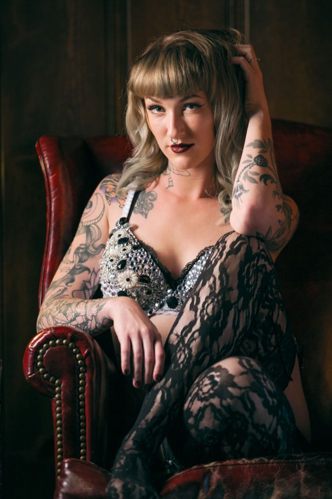 A woman sits confidently in a deep red leather armchair, her pose effortlessly sensual and commanding. Her body is partially obscured by the strategic positioning of her bent leg, clad in sheer black lace stockings, which draws attention to her curves while subtly concealing her torso. She wears an embellished black bra adorned with intricate beading and floral appliqués, adding a touch of glamour and texture. One hand rests gracefully on her thigh, while the other runs through her tousled blonde waves, framing her face with effortless allure. Her tattooed arms, covered in bold floral and geometric designs, become the focal point, enhancing the artistic and edgy aesthetic. The warm, moody lighting casts a soft glow on her skin, complementing the rich tones of the vintage-inspired setting. This pose is a perfect example of boudoir poses that obscure torso, as the combination of her leg placement, arm positioning, and dramatic lighting naturally shifts focus away from the midsection while maintaining an elegant and sultry composition.