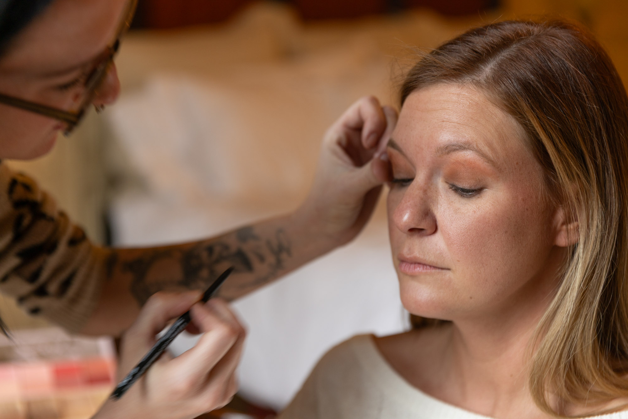 In a close-up shot filled with warm tones and soft focus, a woman with light brown hair and a serene expression sits calmly with her eyes closed while another person, whose tattooed arm is visible, delicately applies eye liner application to her upper eyelid. The makeup artist holds a precise black eyeliner pencil in one hand while gently steadying the client’s eyelid with the other, demonstrating careful control and attention to detail. The woman's skin shows a natural, even texture with a subtle blush and eyeshadow already in place, hinting at a nearly finished look. The background is softly blurred, drawing full attention to the intimate, meticulous act of defining her eyes.