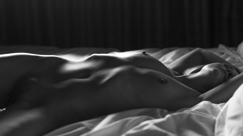 A black and white portrait captures a woman reclining on a softly rumpled bed, her body positioned with graceful symmetry. She lies nude with her arms extended above her head, creating long, elegant lines through her torso and hips. Dramatic side lighting sculpts her form with precise highlights and shadows, emphasizing the contours of her ribs, waist, and legs. Her face is partially obscured in shadow, her eyes closed, conveying a calm and introspective mood. The contrast between the bright white sheets and the dark backdrop adds depth and focus to the composition, resulting in a refined, artistic portrait that blends intimacy and classical styling.