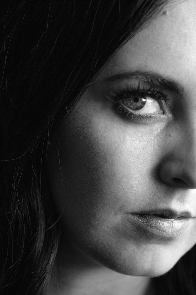 A woman with dark wavy hair gazes toward the camera in this close up black and white portrait from her boudoir photo session. Dramatic side lighting creates deep short light shadows on one half of her face while highlighting her eye and lips in soft grayscale tones against a simple dark background.
