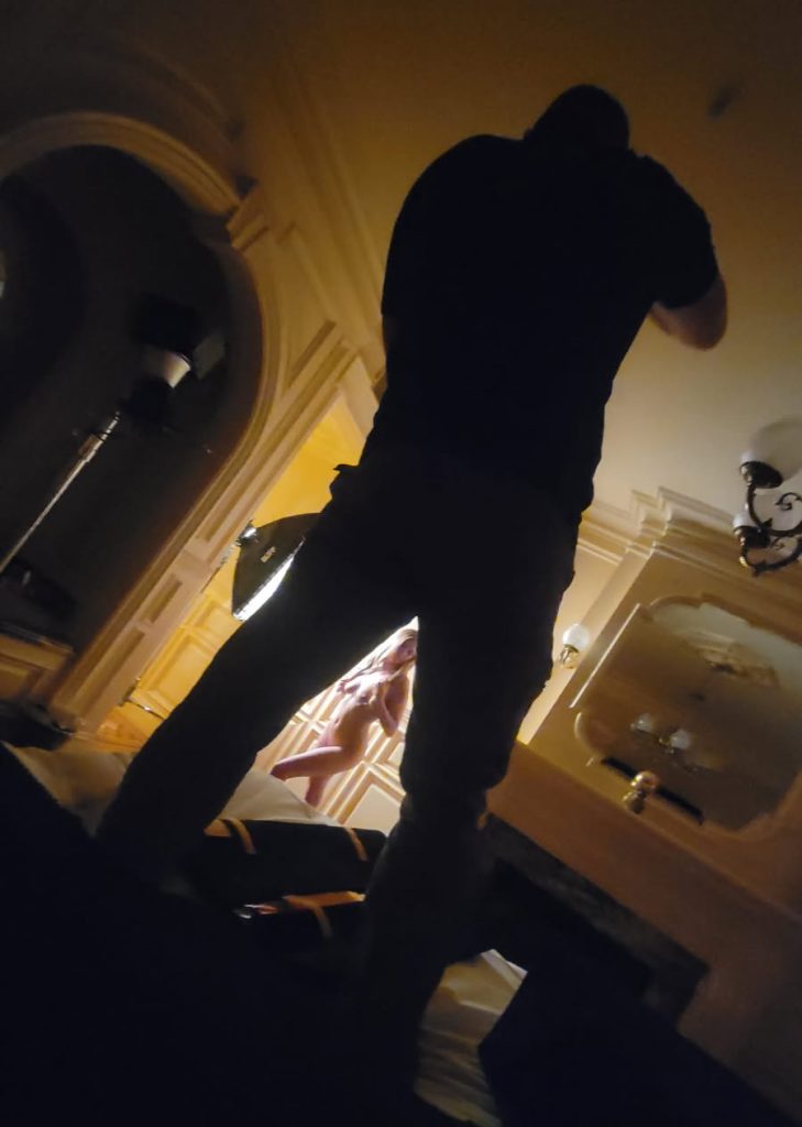 In a richly detailed, low-lit room featuring ornate molding and soft golden illumination, a behind-the-scenes moment captures the atmosphere of boudoir bts with Jason Guy. The image is shot from a dramatic, low angle, with Jason Guy silhouetted in the foreground as he stands poised with his camera, focusing intently on a nude woman who is elegantly posed near a warm light source in the background. Her body is bathed in soft studio lighting from a nearby softbox, highlighting her form against the classic wood-paneled wall. The intimate, artistic setting and the photographer’s commanding presence frame a scene that reveals the technical and creative energy driving a professional boudoir shoot.