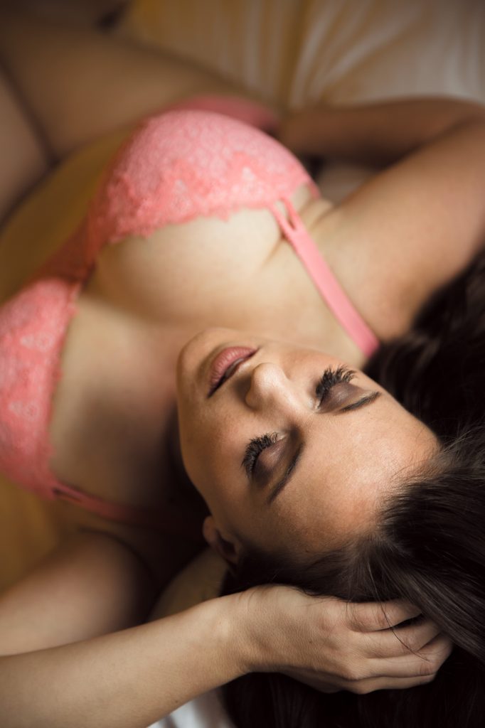 A woman with dark hair lies on her back in a relaxed pose with her eyes closed during a boudoir photography session. She wears a pink lace bra and rests one hand near her hair while soft warm lighting creates gentle shadows across the composition. The high angle view emphasizes the intimate and flattering perspective of curvy boudoir photography.