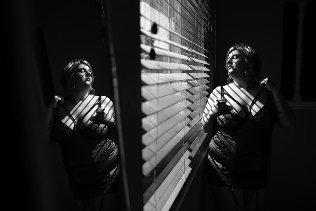 A woman with shoulder-length wavy hair stands near a window with horizontal blinds in a dramatic black and white boudoir photograph. She holds the straps of her black lace slip while strong stripes of light and shadow fall across her body creating lighting and shadows that flatter curvy women. Her eyes are closed and her face is tilted upward as the high contrast composition highlights her form against the dark interior setting.