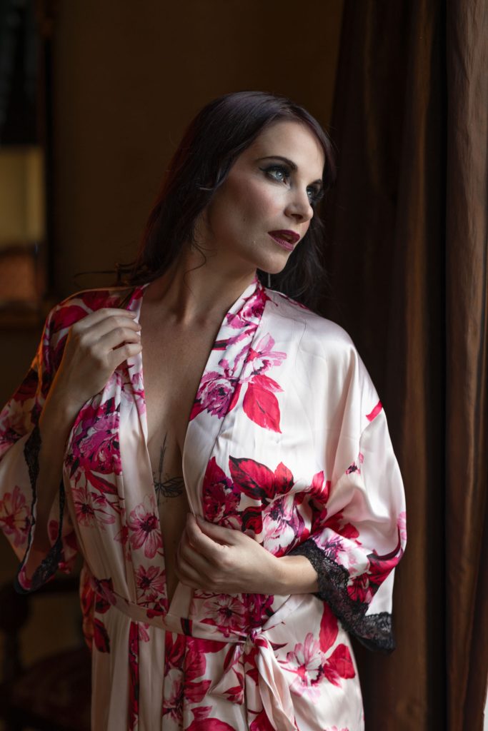 Woman in pink floral silk robe posing by curtained window during her Sonoma County boudoir session, she's cross lit by a large window wearing dark, dramatic makeup with blue eyeshadow