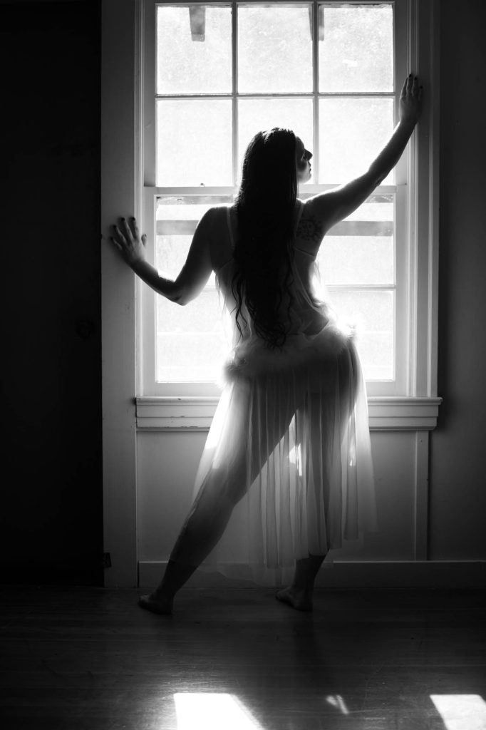 A black-and-white boudoir photograph captures a woman with long wavy hair in silhouette against a bright window, creating strong backlighting and high contrast. She stands in a graceful, elongated pose with one hand pressed to the window frame and the other extended along it, legs in a wide stance that emphasizes elegant lines. She wears a flowing, sheer white babydoll nightgown with thin straps and a layered tulle skirt, one of the best boudoir outfits for women with broad shoulders as the soft, draping fabric and delicate design soften the upper body while highlighting feminine movement and form. The composition uses natural daylight streaming through multi-pane windows to frame her figure dramatically against the dark interior.