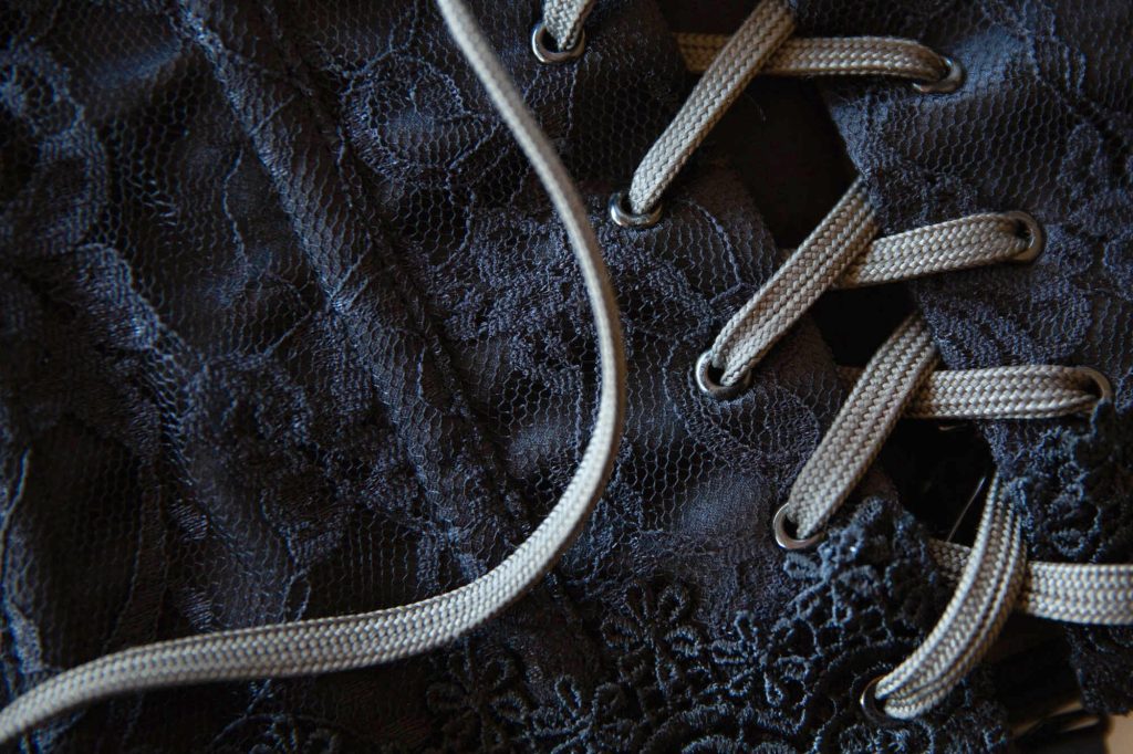 a close-up of a black lace corset. Soft lighting highlights the intricate floral patterns in the dark fabric. Light colored flat laces thread through the metal eyelets to showcase tips for upgrading your corset. Replacing cheap satin lace with quality flat laces like these is an easy and inexpensive way to make a budget corset look more expensive without breaking the bank.