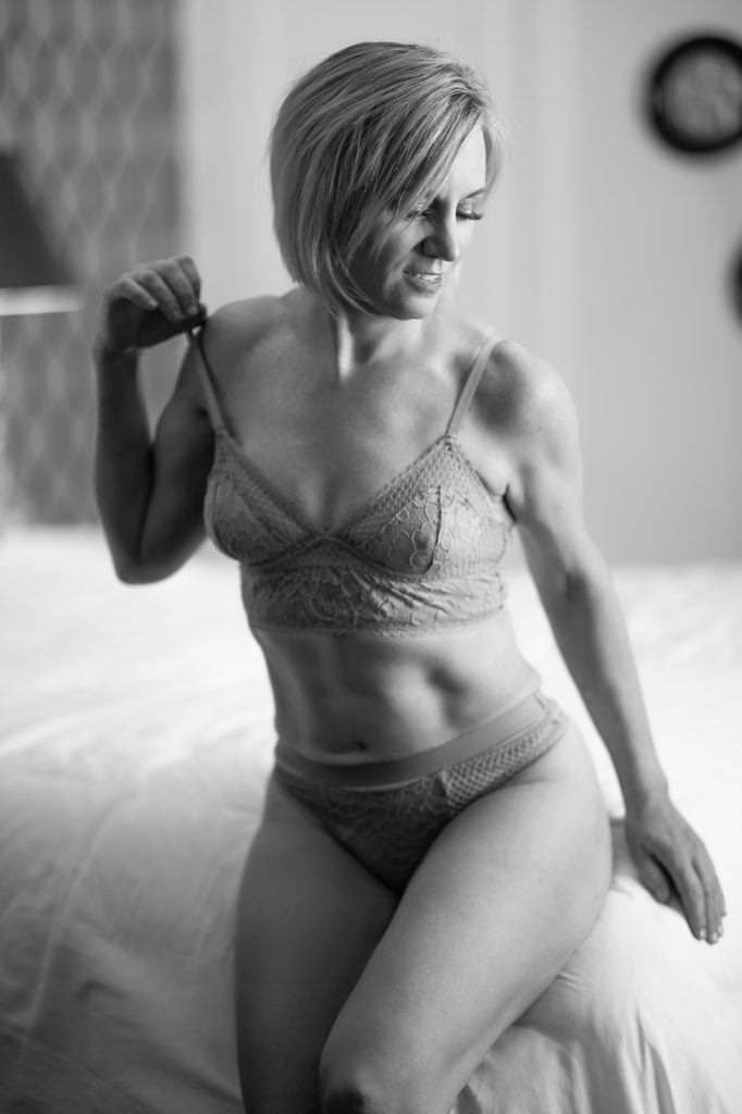50-plus boudoir photos in timeless black and white capturing a confident mature woman with short blonde hair seated on rumpled white bedding, gently adjusting the strap of her delicate lace lingerie with a soft downward smile. Soft diffused lighting highlights her toned physique and natural contours against a blurred neutral background with subtle wall decor.