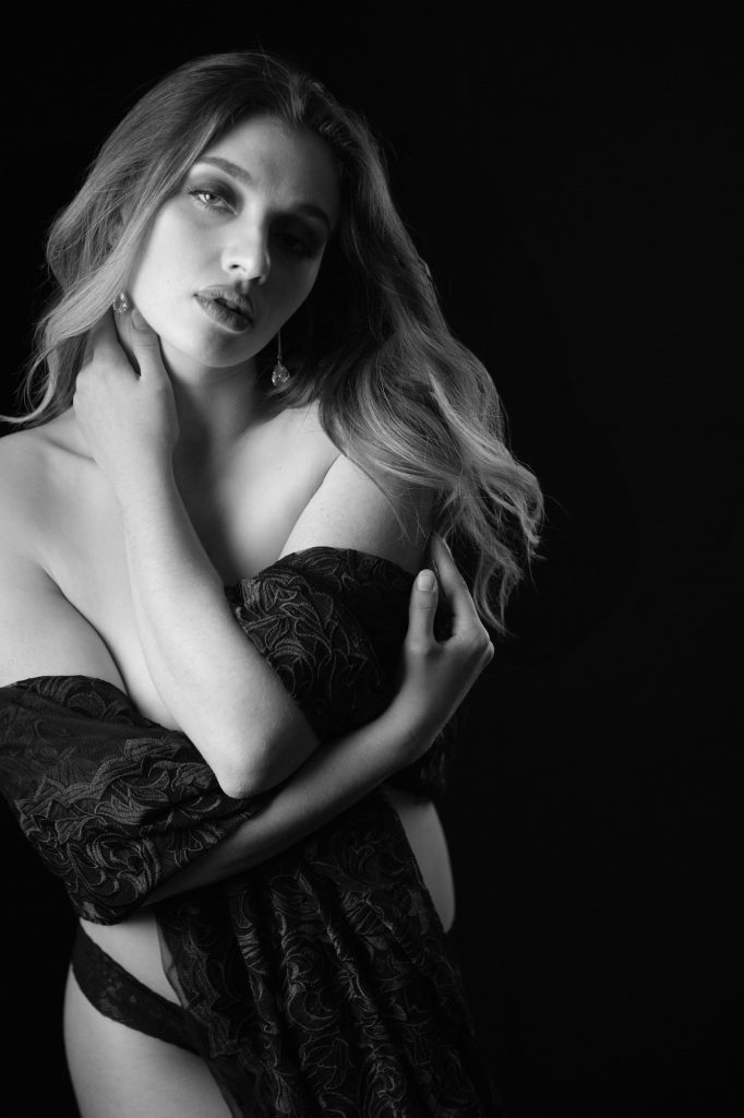 A woman with wavy blonde hair poses elegantly in a black and white portrait for this bay area boudoir photography session, gazing downward with one hand at her neck and the other holding a draped lace fabric across her torso. The composition isolates her against a stark black background, using dramatic low-key lighting to emphasize high-contrast shadows and highlights in monochromatic grays for a timeless, introspective mood.