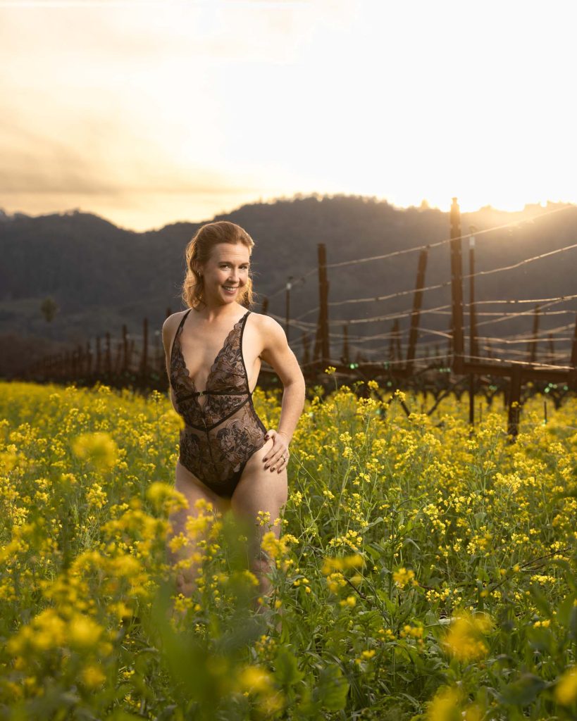 A confident woman in an elegant black lace bodysuit stands with one hand on her hip and a joyful smile, surrounded by vibrant yellow blooming Napa mustard fields with vineyard rows and silhouetted hills in the background, bathed in warm golden sunset light during a boudoir photoshoot in Napa mustard fields.