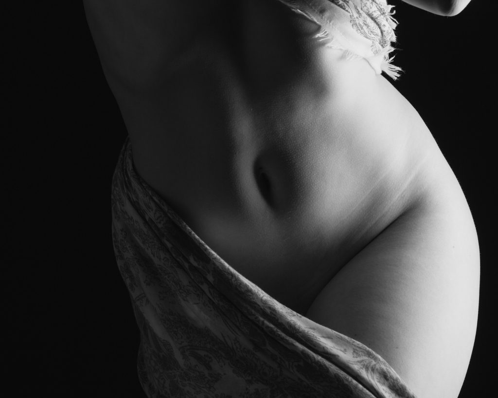 A woman poses in this black and white close-up photograph that demonstrates the kind of photos you can get if you follow these boudoir photo shoot tips. This photo features dramatic lighting and artistic composition. Strong side lighting creates bright highlights and deep shadows across her abdomen and hips. Patterned fabric with frayed edges drapes around her lower and upper torso exposing her belly button. The shadows fade into the black background like she's coming out of the shadows. 