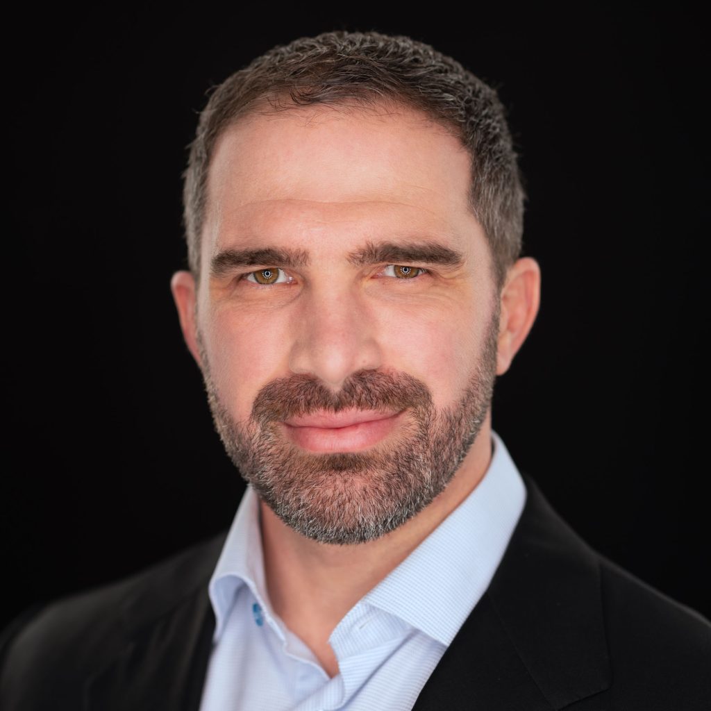 A professional headshot of a man with short, dark hair and a well-groomed beard. He has a confident yet approachable expression with a slight smile. His eyes are sharp and focused, framed by thick eyebrows. He is wearing a crisp light blue dress shirt and a black blazer. The background is solid black, creating a striking contrast that highlights his facial features. The lighting is even and well-balanced, emphasizing the texture of his beard and the details of his skin. His posture is relaxed but assured, giving a strong, professional presence.