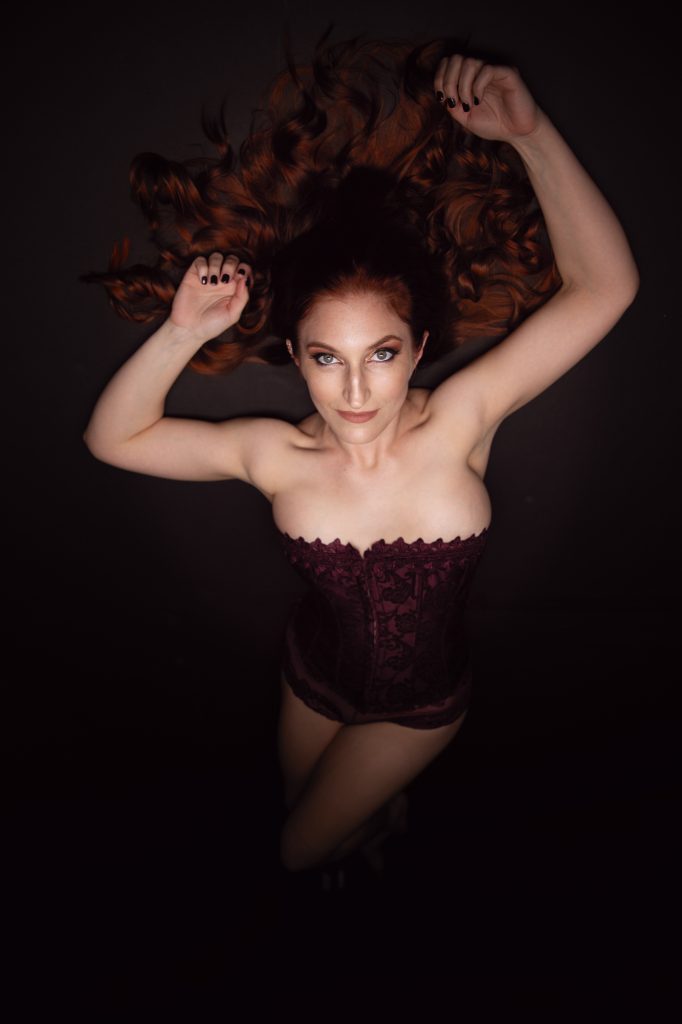 Boudoir photography Santa Rosa image capturing a woman with long wavy auburn hair reclining on a dark surface, viewed from above with arms raised holding her hair and gaze toward the camera, attired in a deep maroon lace corset, under low-key lighting that emphasizes rich earthy tones and dramatic shadows for a composed atmosphere.