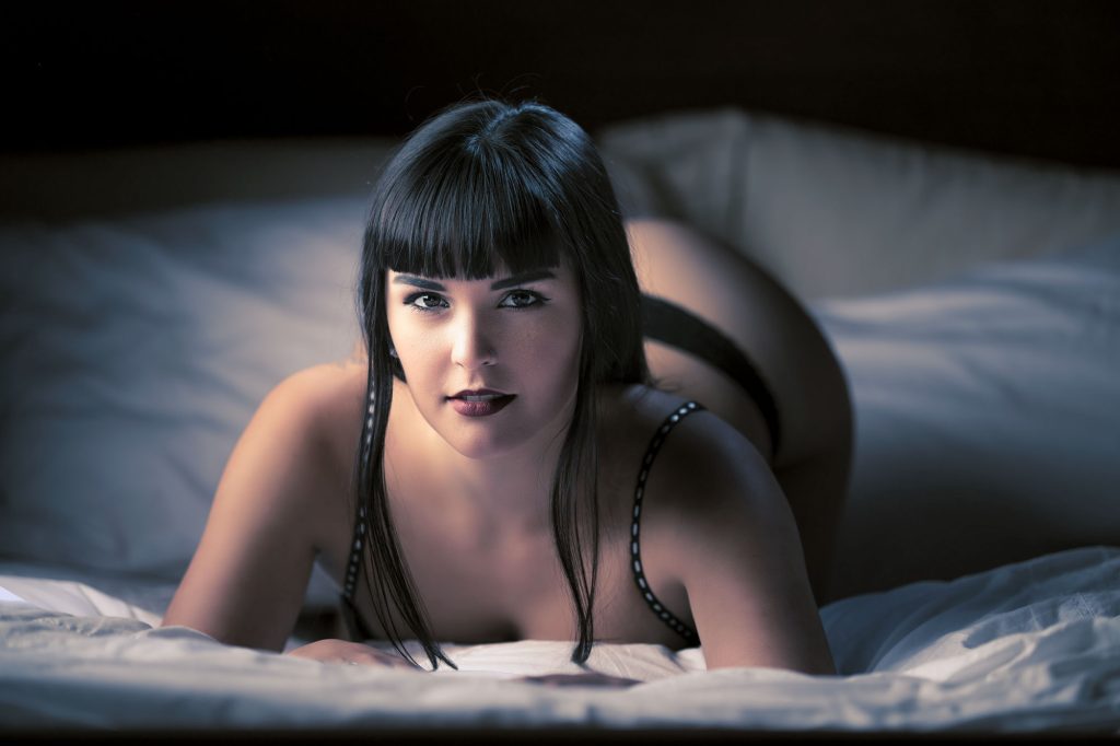 A woman with straight dark hair and blunt bangs lies on her stomach atop soft white bedding in this boudoir photos Rohnert Park session, gazing directly at the viewer with a subtle expression, one arm bent to support her chin. The composition emphasizes her upper form against a dimly lit background, using soft blue-tinted lighting to create gentle shadows and highlights in tones of cool grays, blacks, and whites for a serene, intimate atmosphere, while she wears a simple black strap outfit.