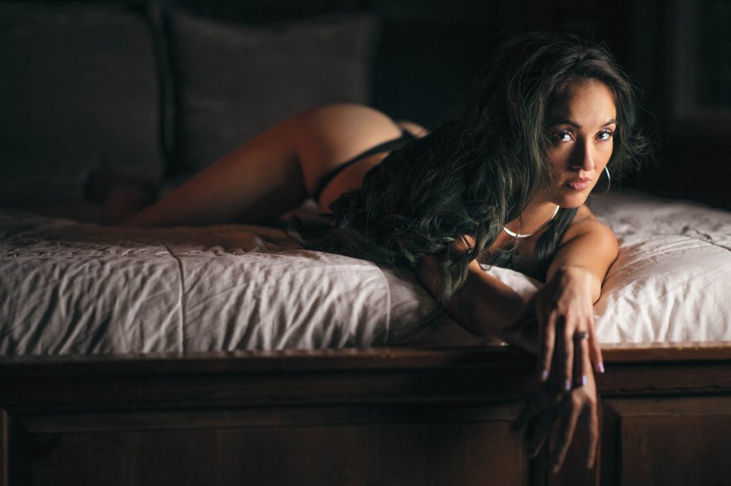 A boudoir photo from Sebastopol showcasing a woman with long dark wavy hair lying relaxed on her stomach atop white rumpled sheets, gazing directly over her shoulder with a subtle expression, wearing thin black strappy top and bottoms, composed with warm side lighting creating golden hues and deep shadows against soft pillows and dark wooden bed frame in a cozy dim room.