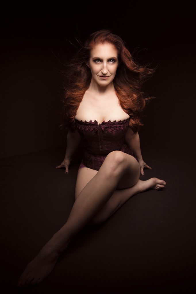 A woman with flowing red hair sits gracefully on the floor in this boudoir portraits in Sonoma session, posed with legs extended to one side and hands supporting her, wearing a deep purple lace corset while gazing directly at the viewer. The composition centers her against a dark backdrop, using dramatic low-key lighting to create warm highlights and deep shadows in tones of maroon, auburn, and black for a confident, intimate mood.