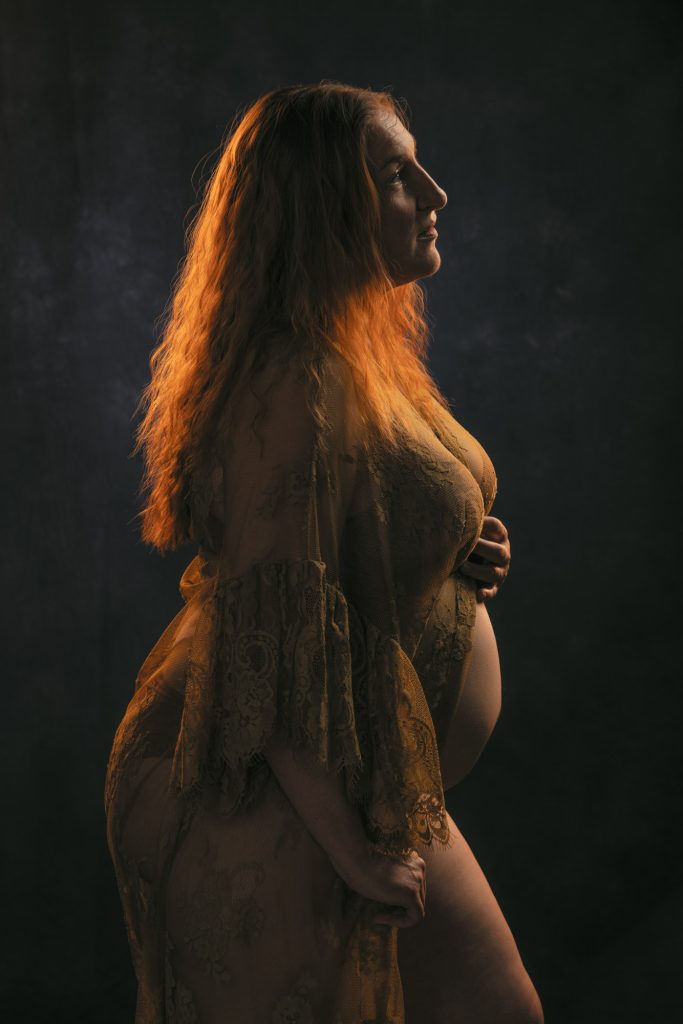 A chiaroscuro pregnancy boudoir photo of a woman with long wavy red hair standing in profile, hand gently cradling her belly in serene reflection, attired in a sheer golden lace gown with flared sleeves, composed with dramatic side lighting casting warm amber highlights and deep shadows against a dark neutral backdrop for a peaceful intimate mood.
