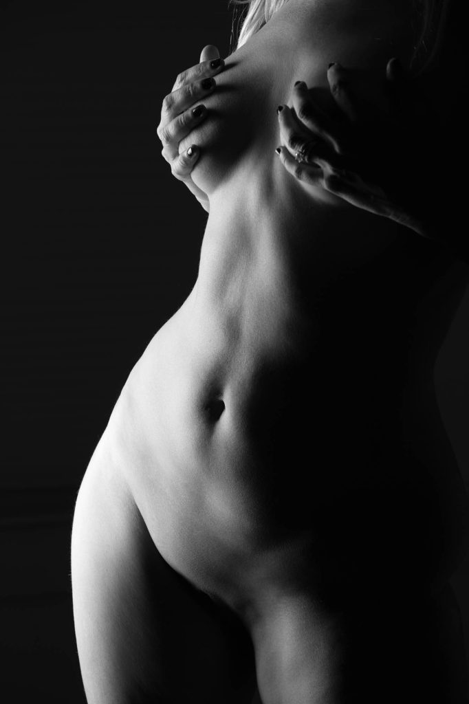 Fine art figure photography in dramatic black and white presenting a close-up side view of a woman's torso, arms gracefully crossed with hands modestly covering her chest and fingers showing dark nail polish and a ring. Strong low-key lighting creates deep shadows and luminous highlights that sculpt the smooth contours and curves against a seamless dark background.