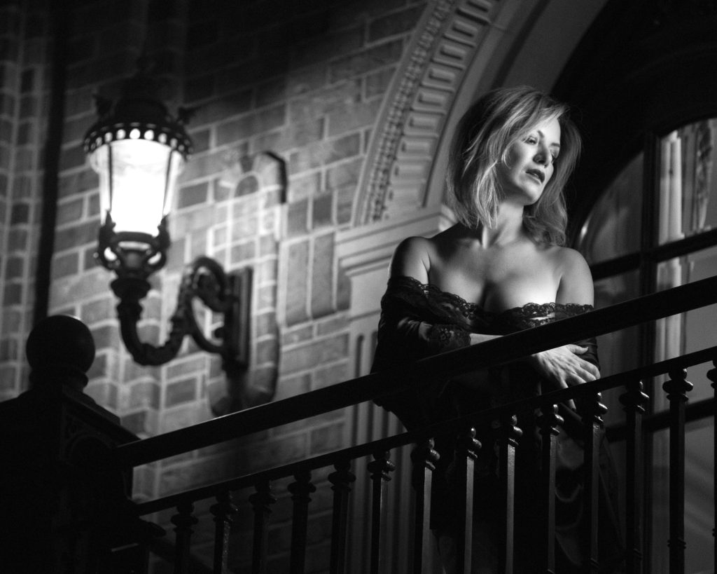 Gothic boudoir photo in black and white showing a woman with shoulder-length blonde hair leaning on a wrought-iron staircase railing in a brick-arched hallway, her pose contemplative with arms crossed and gaze directed away, attired in an off-shoulder lace top with a draped jacket, under dramatic side lighting from a wall lantern that creates deep shadows and high-contrast grayscale tones for a moody architectural composition.