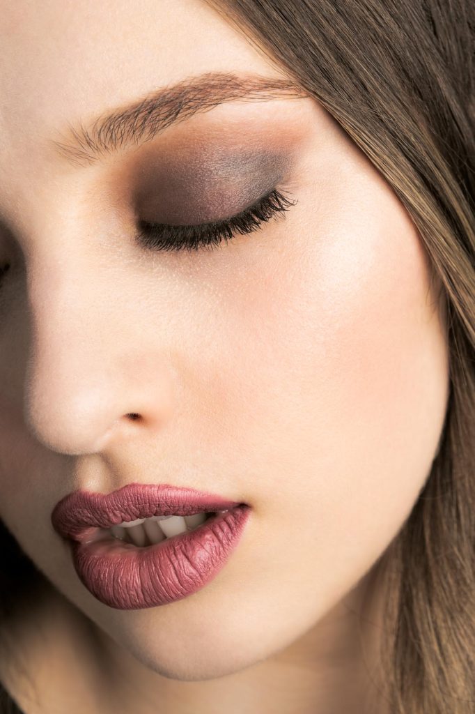A woman with brown hair poses in this close-up portrait photograph that demonstrates what your photos could look like if you follow these lash extension tips for boudoir photos. Gentle highlights accent her smooth skin as she tilts her face slightly with one eye closed to showcase long lashes and neutral smoky eyeshadow while her lips part to reveal matte mauve lipstick against a blurred neutral background.