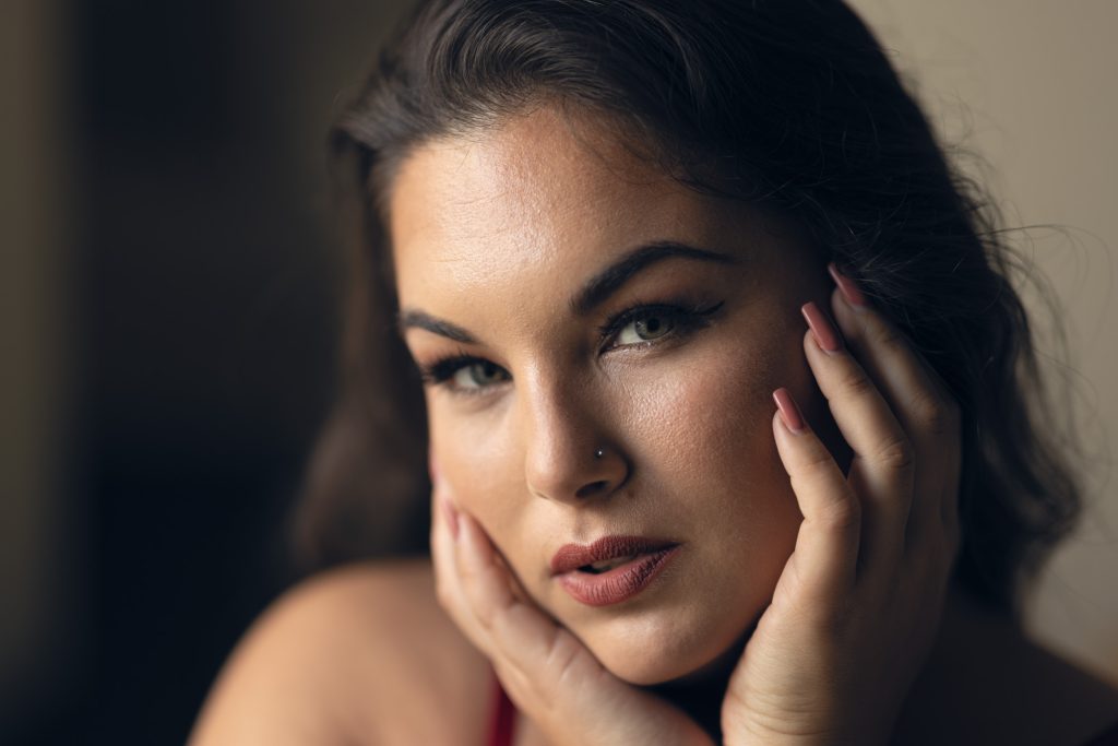 A woman with dark brown hair poses in this close-up portrait that demonstrates the kind of photos you can get if you follow these manicure tips for boudoir photo shoots. She rests her hands with manicured nails on her cheeks and looks directly at the camera. She has green eyes and a subtle nose ring with a red strap visible on her shoulder against a blurred neutral background.