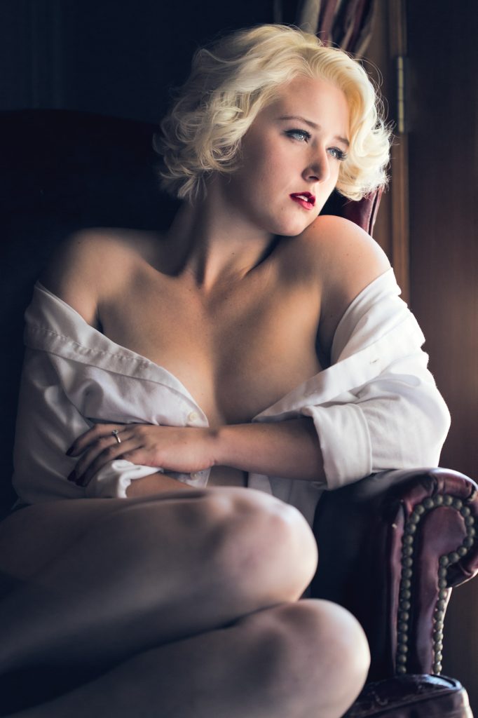 Maralin manroe inspired boudoir photo of a woman with short platinum blonde curls and bold red lips, seated sideways in a deep red armchair with legs drawn up, one hand adjusting a loosely draped white shirt. Soft, directional lighting creates warm highlights on skin and fabric amid dark wooden tones and curtained background, forming a composed, nostalgic scene.