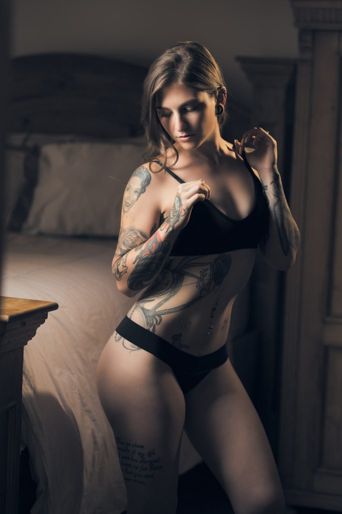 An intimate boudoir portrait by a Marin County photographer features a tattooed woman in black lingerie standing beside a rustic wooden bed. She gently pulls at the strap of her bralette with a serene, introspective expression. Her toned body is adorned with intricate tattoos, including a portrait on her shoulder and script along her thigh. Multiple piercings on her navel and stretched earlobe jewelry add to her edgy style. Warm, directional lighting casts soft shadows that highlight her figure’s contours, enhancing the depth and emotion of the image. The cozy bedroom with plush bedding and vintage furniture contrasts with her bold, artistic presence.