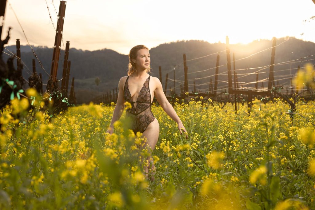 A confident woman in an elegant black lace bodysuit strolls gracefully through vibrant blooming Napa mustard flowers, gazing softly over her shoulder with a subtle smile, framed by vineyard rows and rolling hills under warm golden sunset backlighting with lens flare, captured by a Napa mustard flowers boudoir photographer.