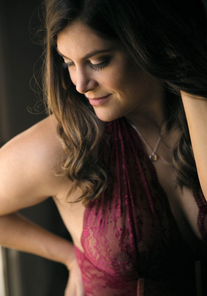 Postpartum boudoir photography image portraying a woman with long wavy brunette hair indoors against a dark background, captured in close-up side profile with a gentle downward gaze and subtle smile, one hand raised near her shoulder, attired in a deep purple lace halter top and necklace, under soft low-key lighting that highlights warm skin tones and creates subtle shadows for a peaceful composition.