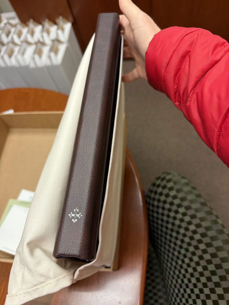 Publishers Mark on JG Boudoir Album: close-up of brown leather spine with ornate silver emblem held by hand in red jacket under soft lighting.