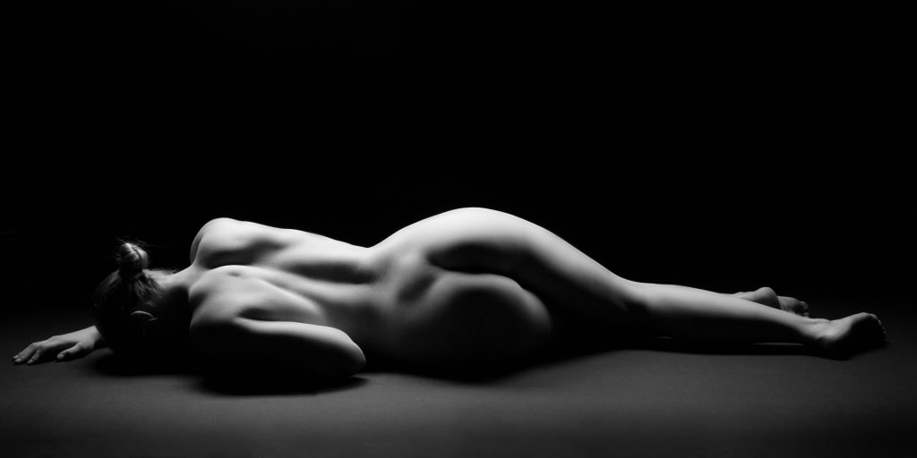 A woman with hair tied in a bun lies prone on a dark surface against a pitch-black background, her arms extended forward and legs stretched out in relaxed repose, captured in high-contrast black-and-white with dramatic lighting sculpting smooth contours and deep shadows along her back and form, an example of sculptural boudoir photos.