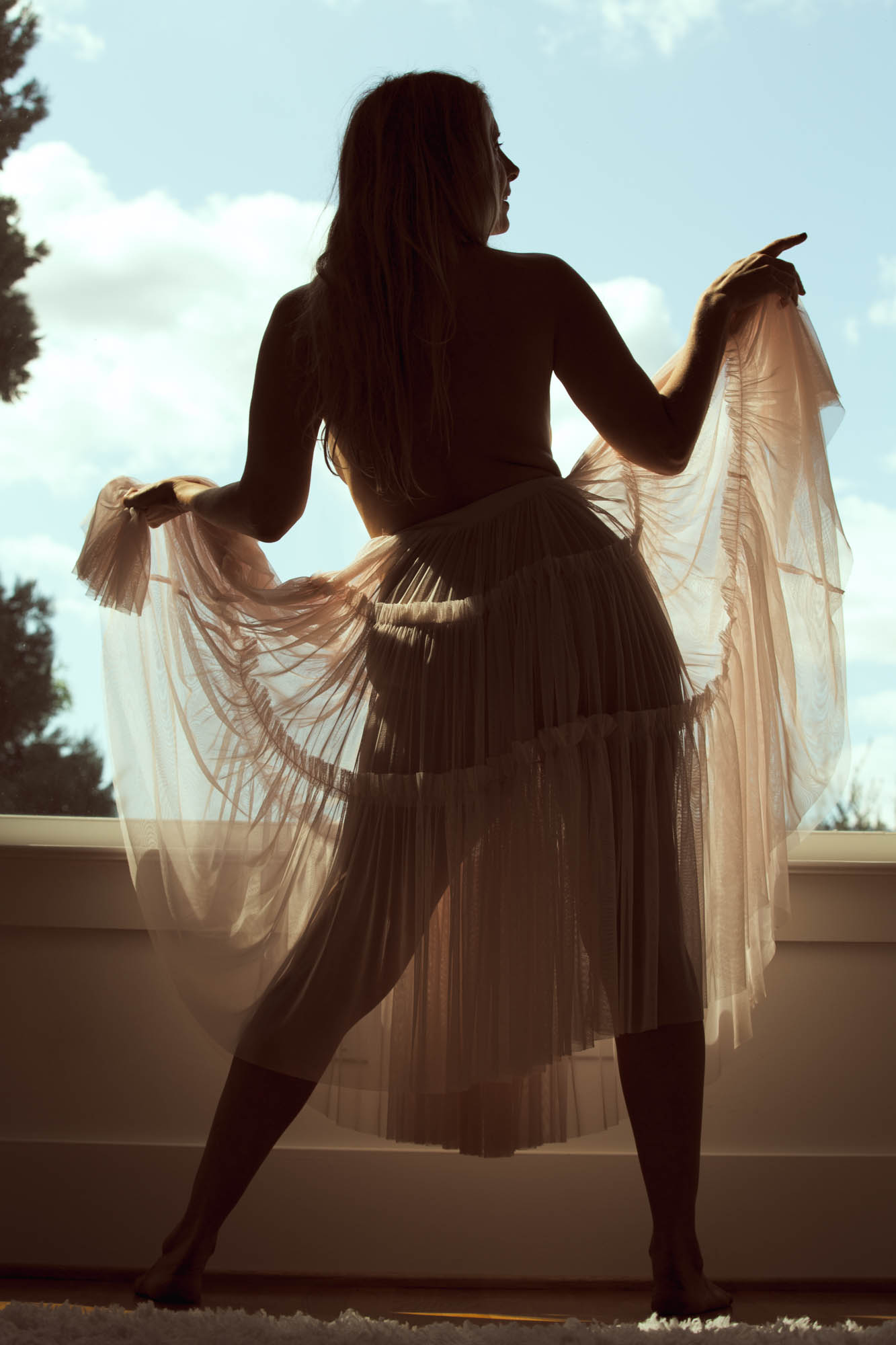 Silhouette tulle skirt boudoir photo depicting a woman in a dynamic back view pose against a bright blue sky with clouds, long hair flowing down her bare back as she extends arms to hold sheer light-colored tulle skirt fabric outward, one leg stepped forward barefoot on a light floor; strong backlighting creates a dark silhouette effect with warm golden edges and subtle transparency in the skirt, contrasted by soft outdoor greens and blues for an intimate composition.